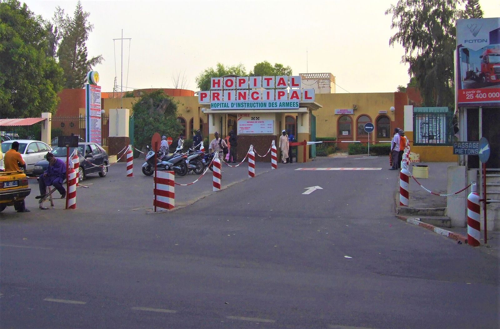 Entrée de l’Hôpital Principal de Dakar, avec son portail, des barrières rouges et blanches et des passants