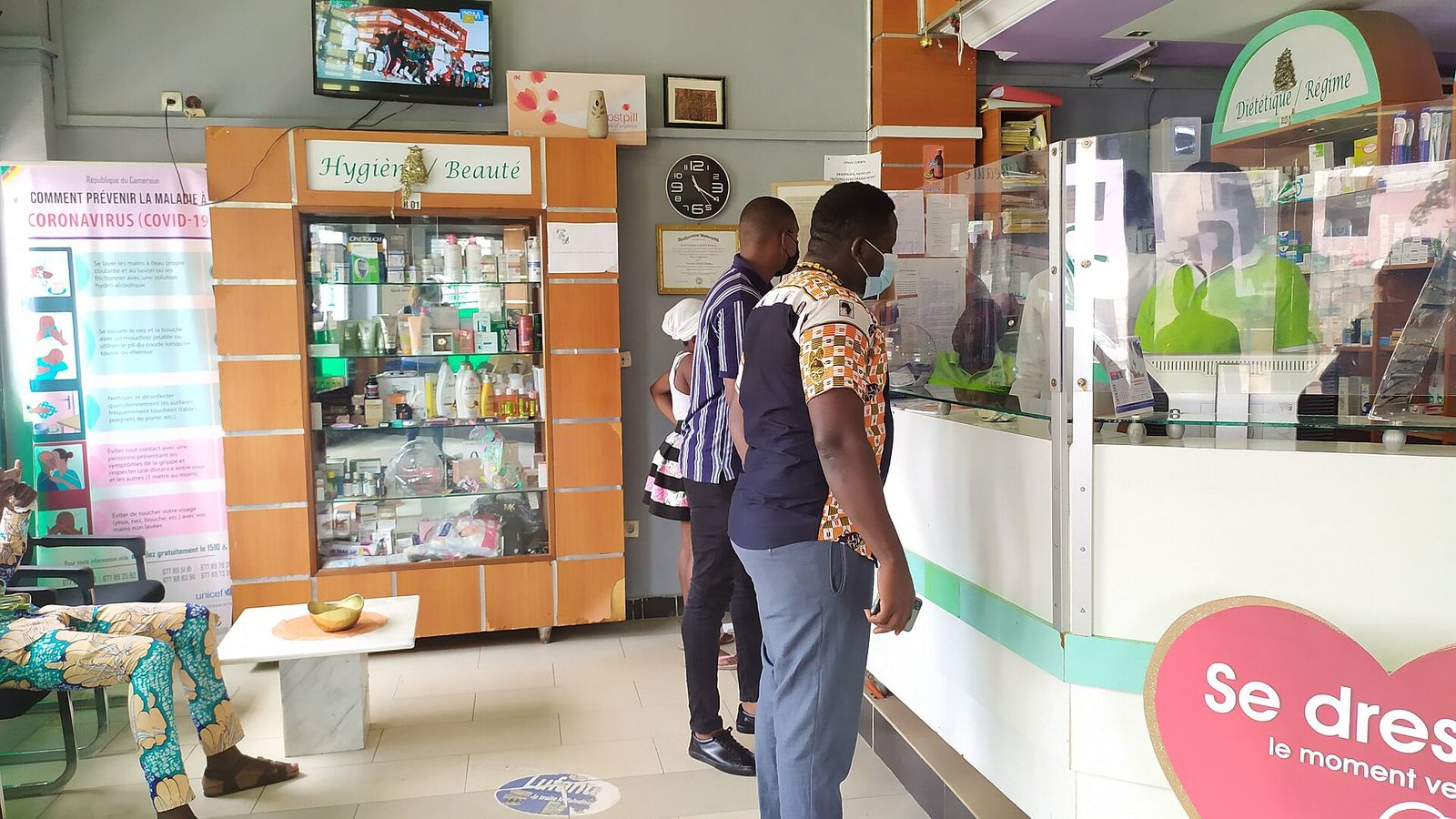 Deux clients au comptoir d’une pharmacie moderne au Cameroun, devant des rayons de produits d’hygiène et de beauté
