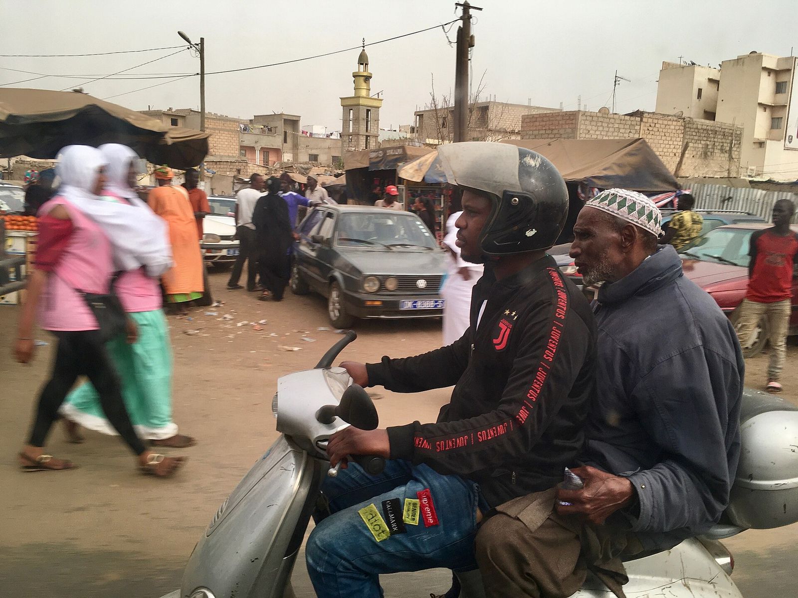 Un homme âgé coiffé d’un bonnet blanc, passager d’un scooter dans une rue animée d’une ville du Sénégal